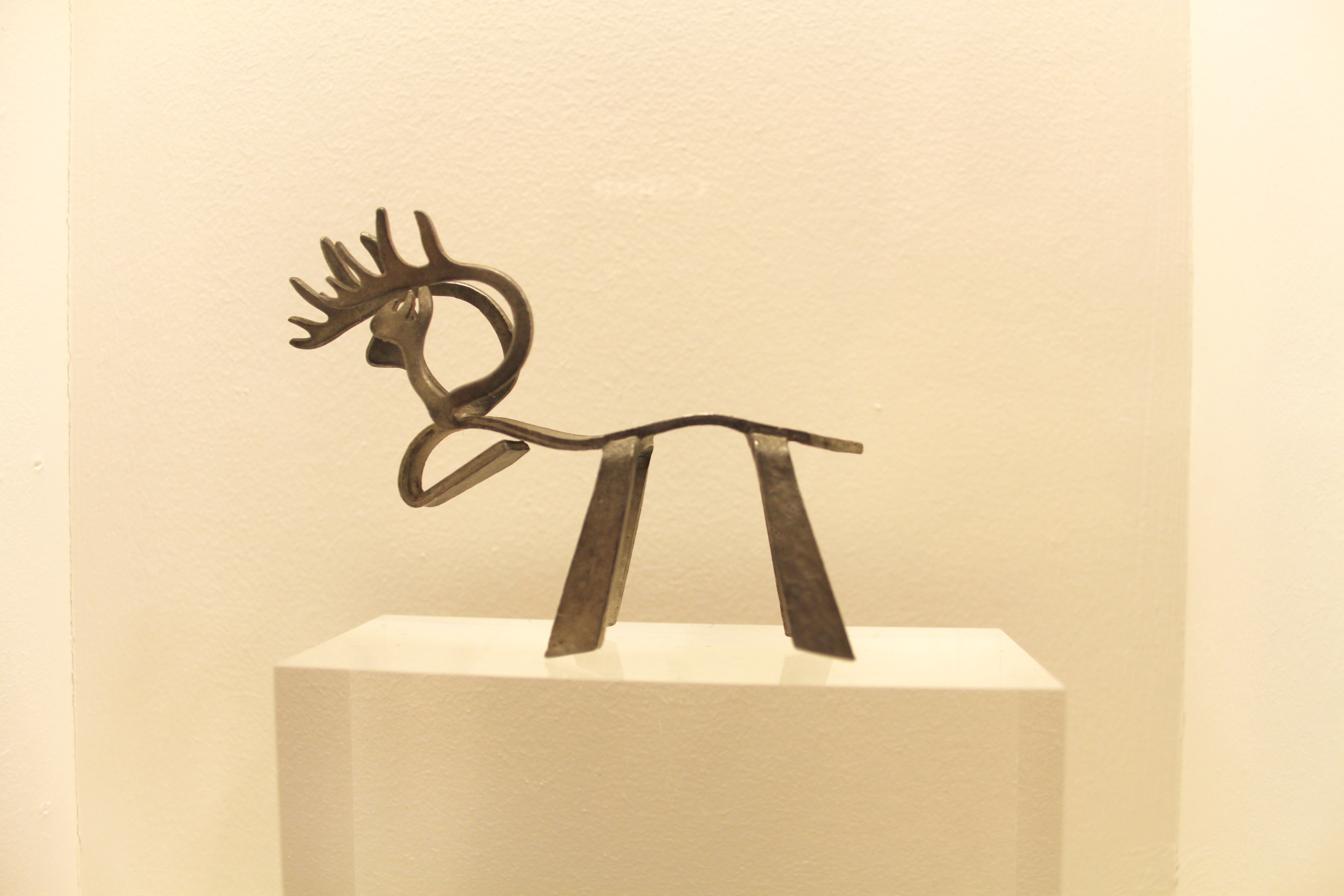 A small artistic elegant metal reindeer with a curved back and head, simple legs and intricate antlers  on a small off-white podium with an off-white wall behind.
