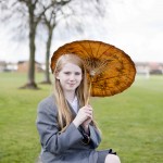 Pupil in uniform holds pretty autumnal-coloured parasol in park with trees behind.