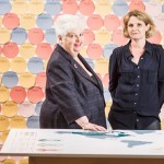 Barbara and Sophie, both dressed in black, stand between a table with a map of the world and a wall of coloured (blue, yellow, red and pink) discs on which exhibition visitors have written their own or their family's migration or non-migration stories.